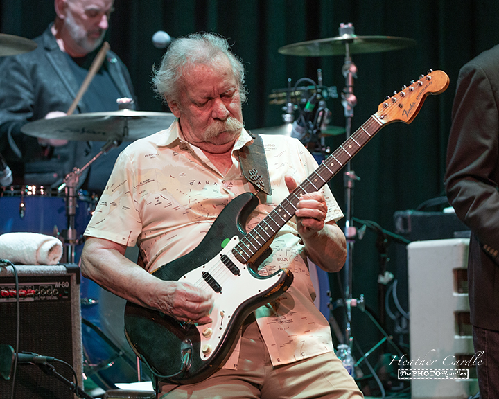 A man playing an electric guitar on stage with a set of drums in the background