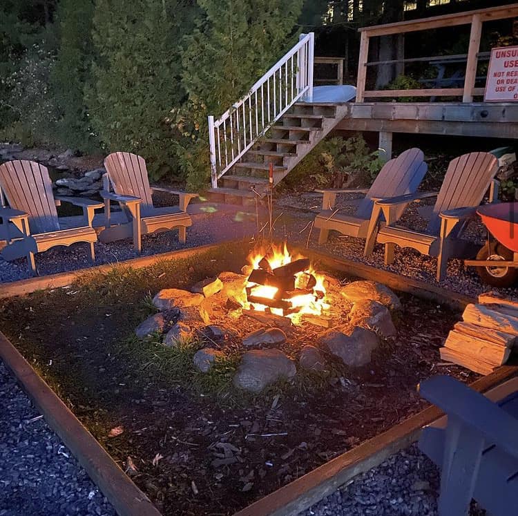 A campfire surrounded by some nice Highlands chairs in front of stairs and a deck on a waterfront