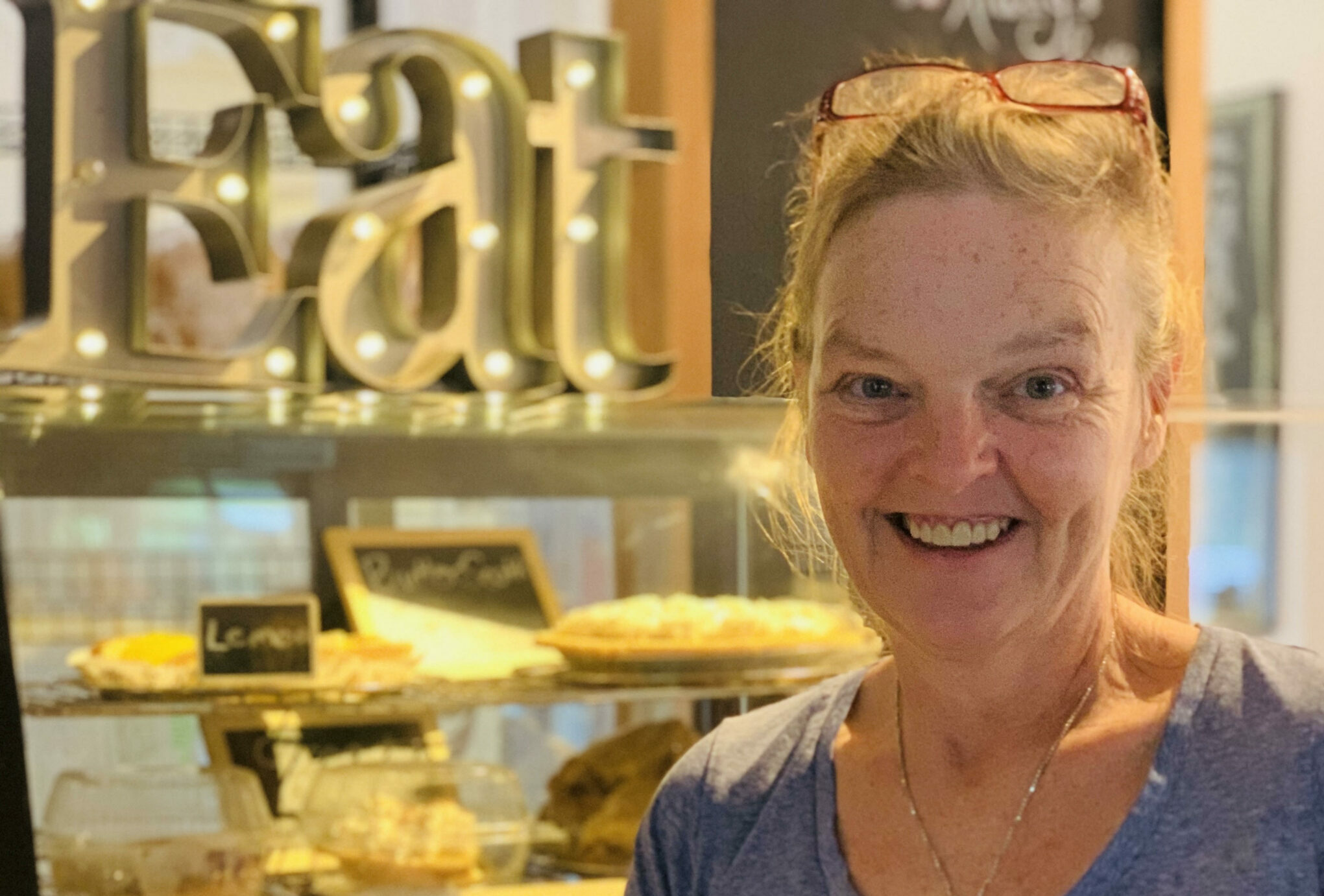 Owner Maggie of the Rustic Roadside Restaurant smiling at the camer with the word EAT and baked goods in the background